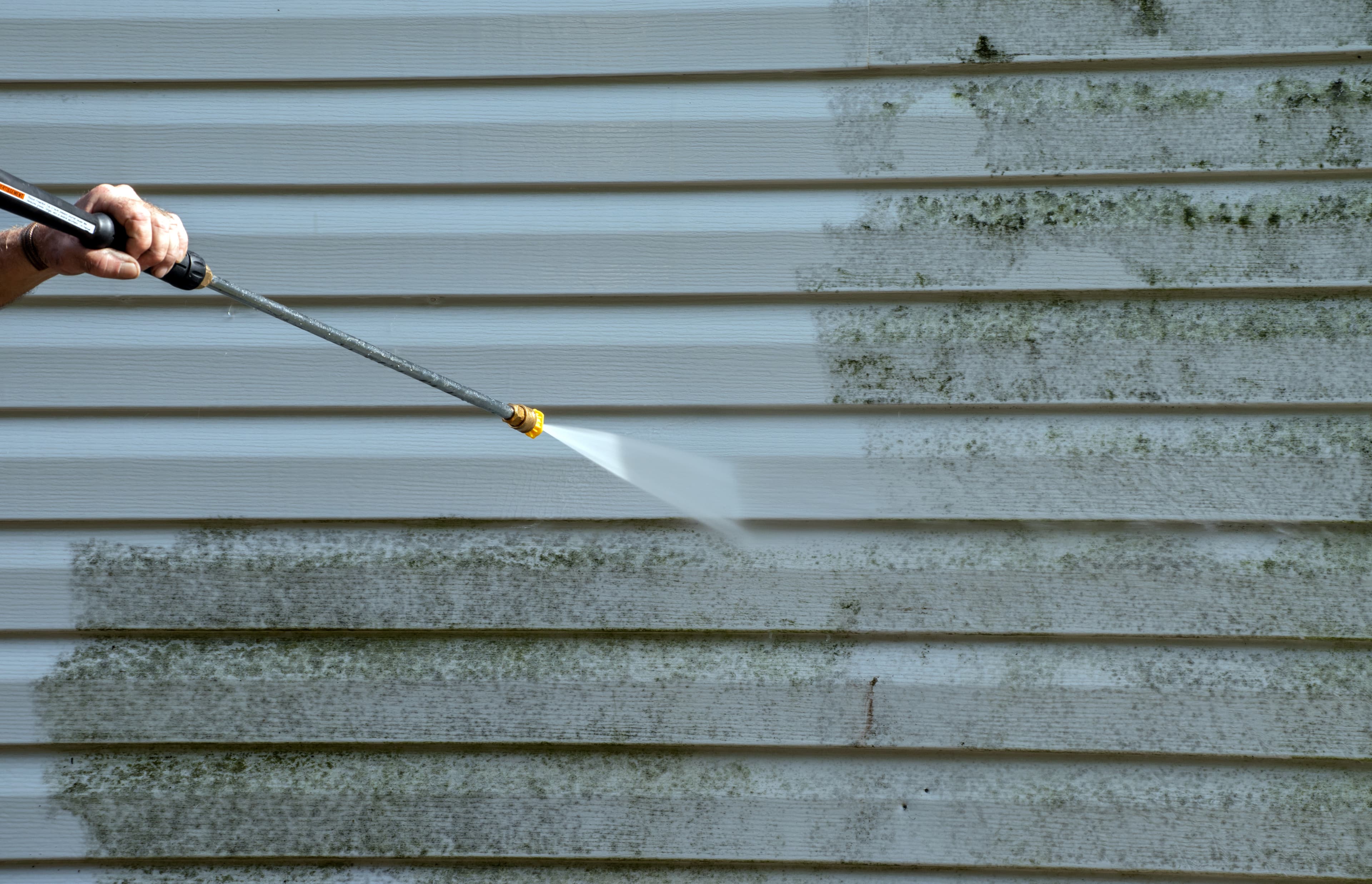 A person pressure washing the siding of a home which has dark green moss being cleaned off of it.