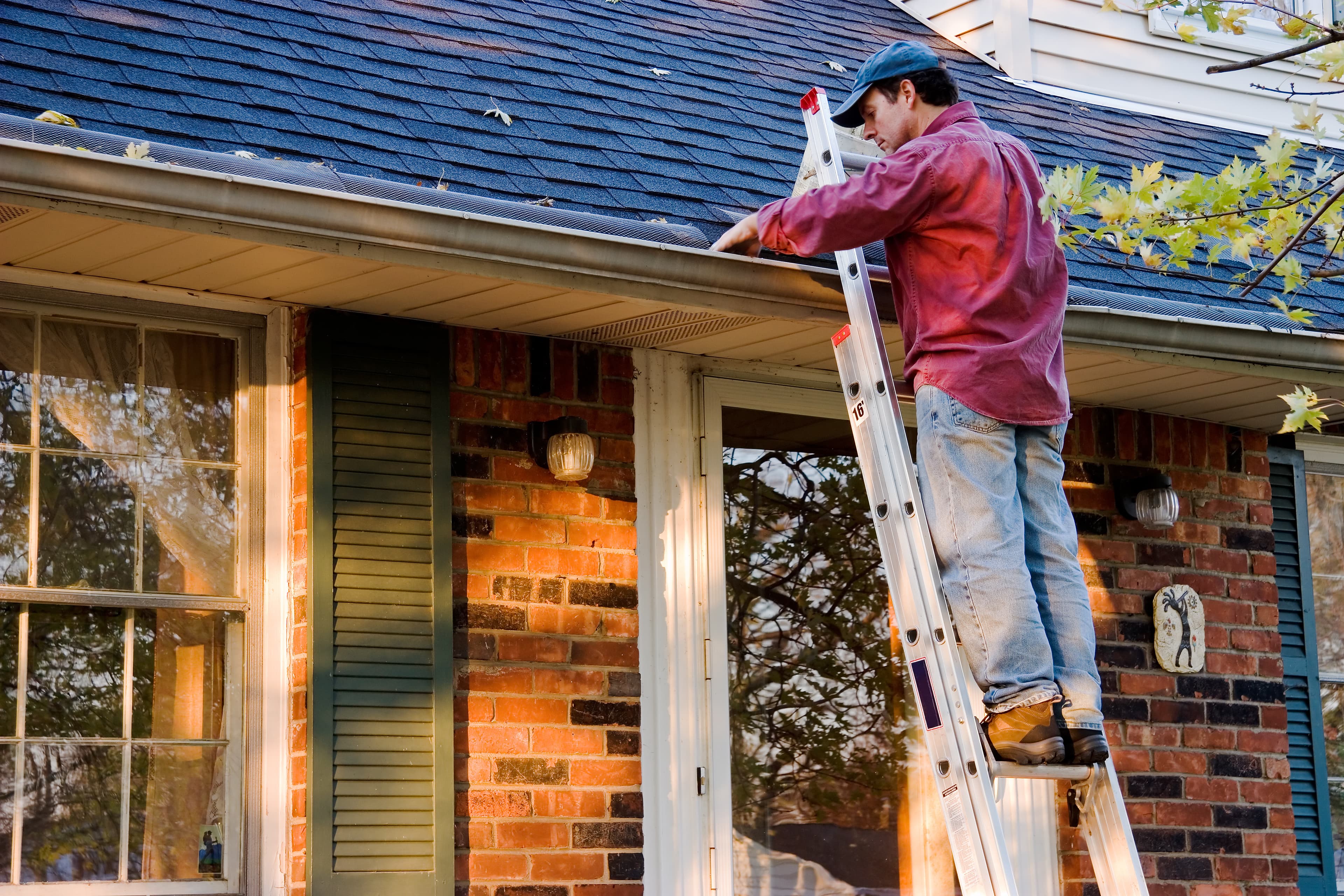 man in maroon shirt on a ladder removing debris from gutters on a house with green shutters